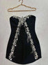 Vestido Mini preto sem alças, em tecido liso, adornado com flores de cristais prateados e correntes brilhantes